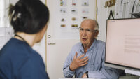 Bildet viser en sykepleier som sitter ved pc-en og snakker med en eldre, mannlig pasient