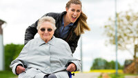 Bildet viser en eldre dame i rullestol som er ute på tur med en ung kvinne.