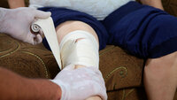 Bildet viser hendene til en sykepleier som bandasjerer et kne til en pasient hjemme i stolen sin.