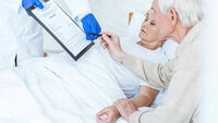 Bildet viser en syk kvinne i senga på sykehuset som har besøk av sin mann. En lege gir dem et skjema og en penn.