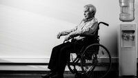 Bildet viser en eldre mann som sitter i en rullestol ved siden av en vanndispenser